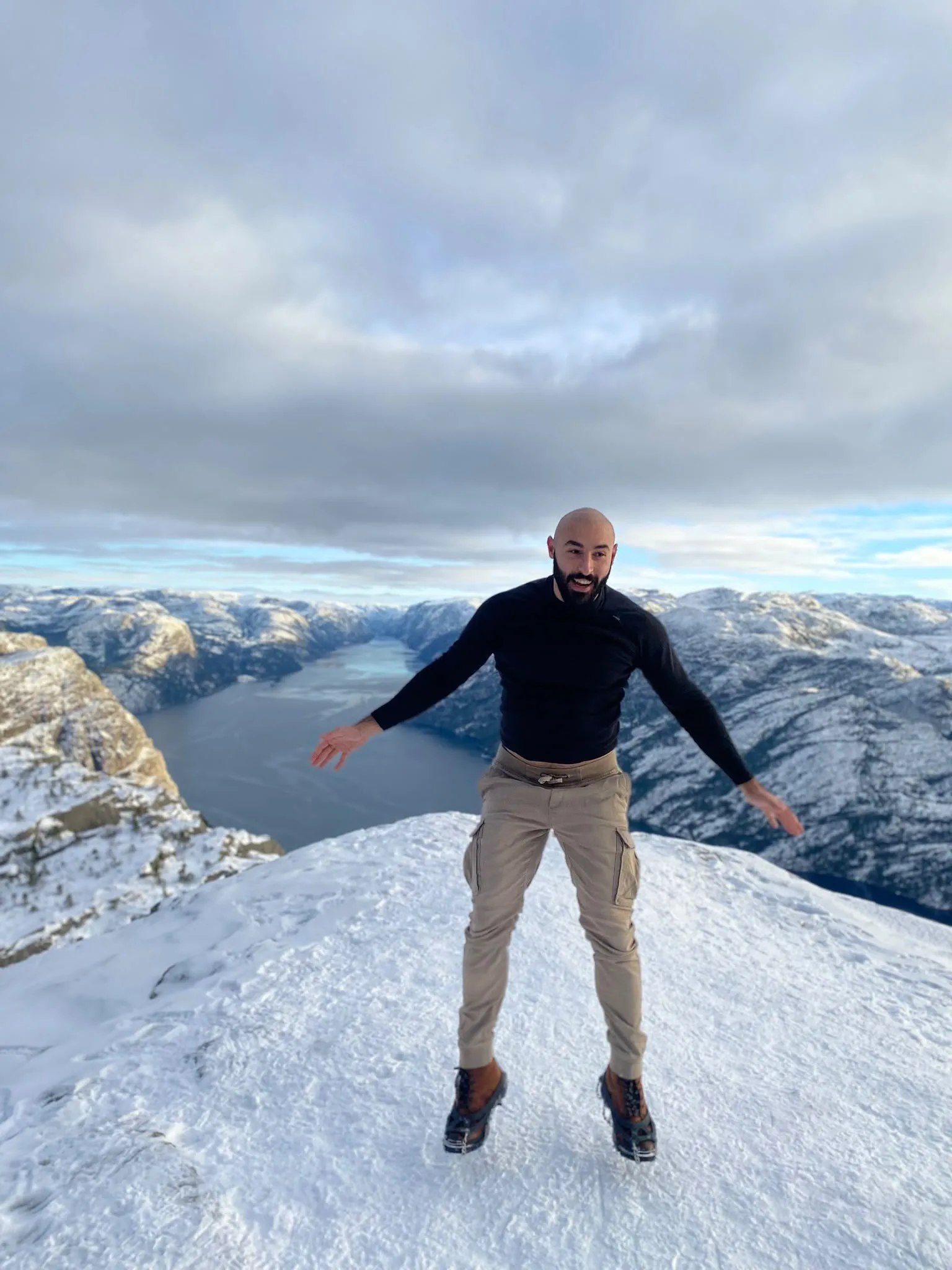 Daniel staat met gespreide armen op een besneeuwde bergtop boven een fjord